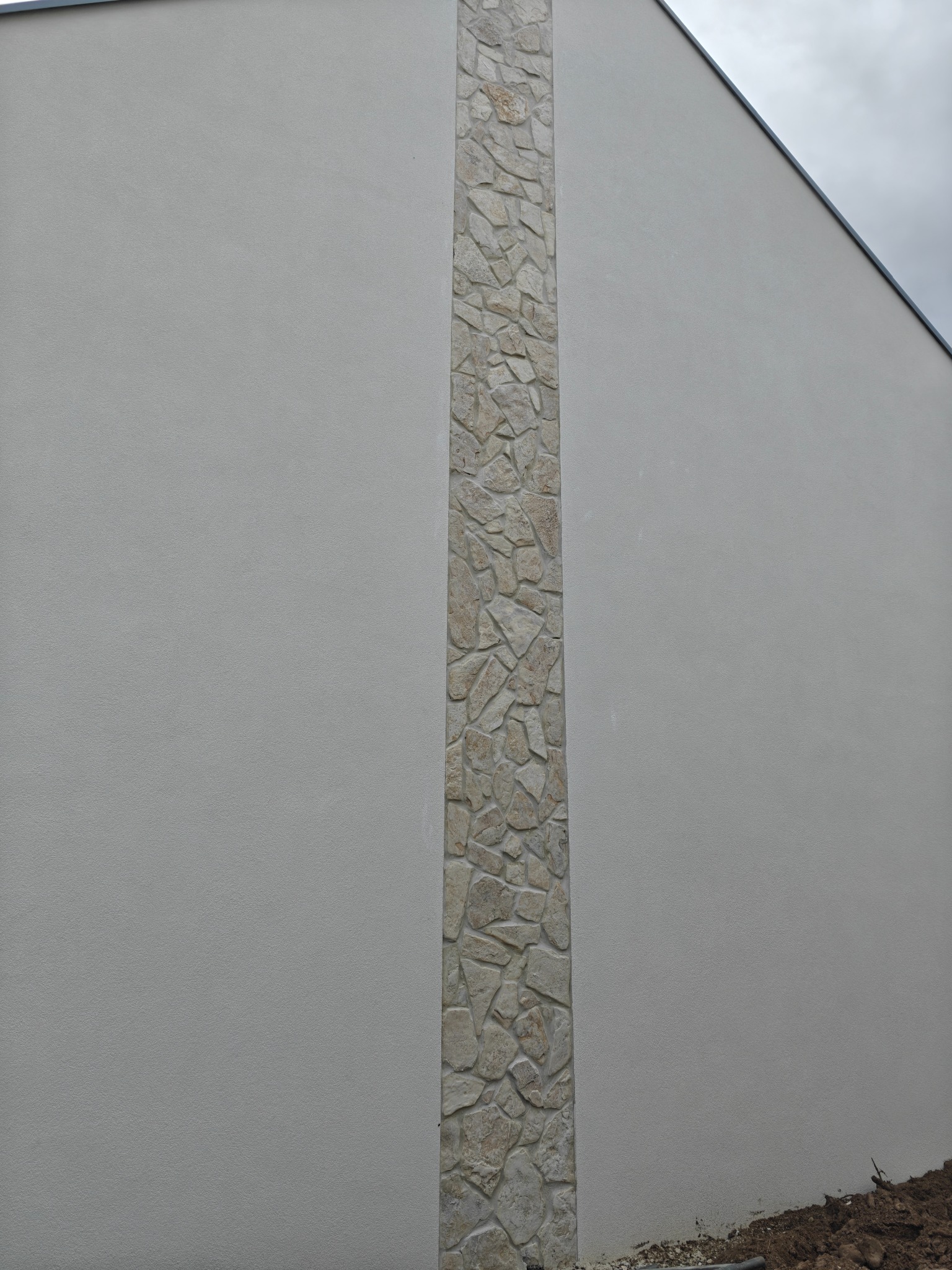 Szara elewacja budynku z pionowym pasem dekoracyjnym z kamienia naturalnego. Widok z dołu, pochmurne niebo w tle. Fragment ziemi u dołu kadru.