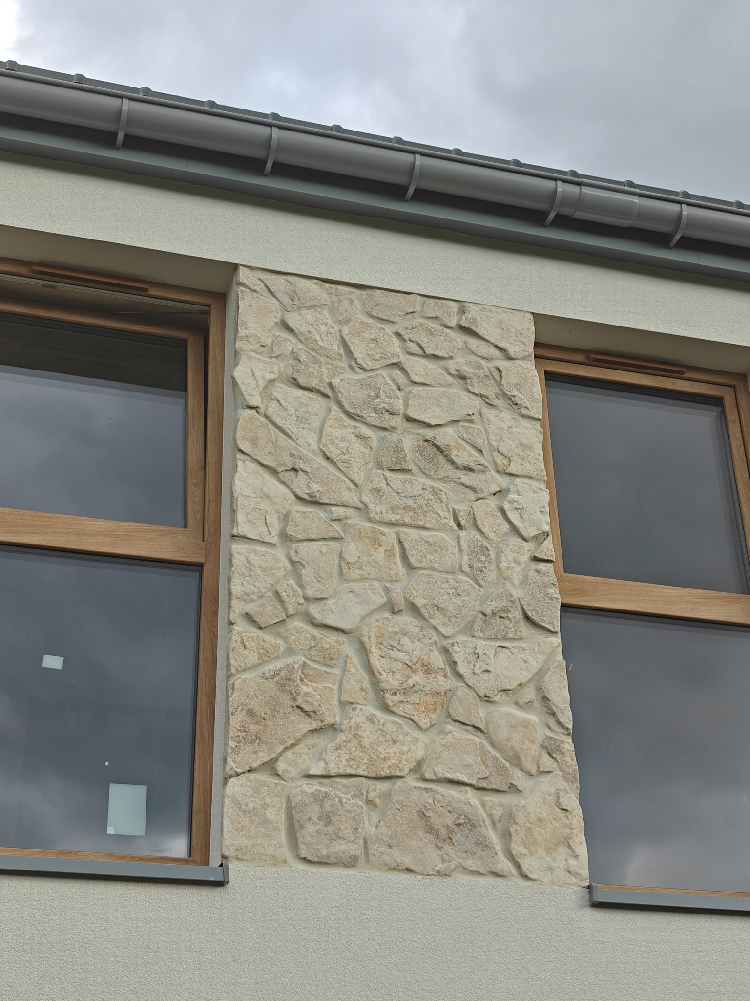 Fragment elewacji budynku z oknami i dekoracyjnym panelem z kamienia naturalnego w odcieniach beżu. Widoczne szare rynny i fragment elewacji o fakturze baranka.