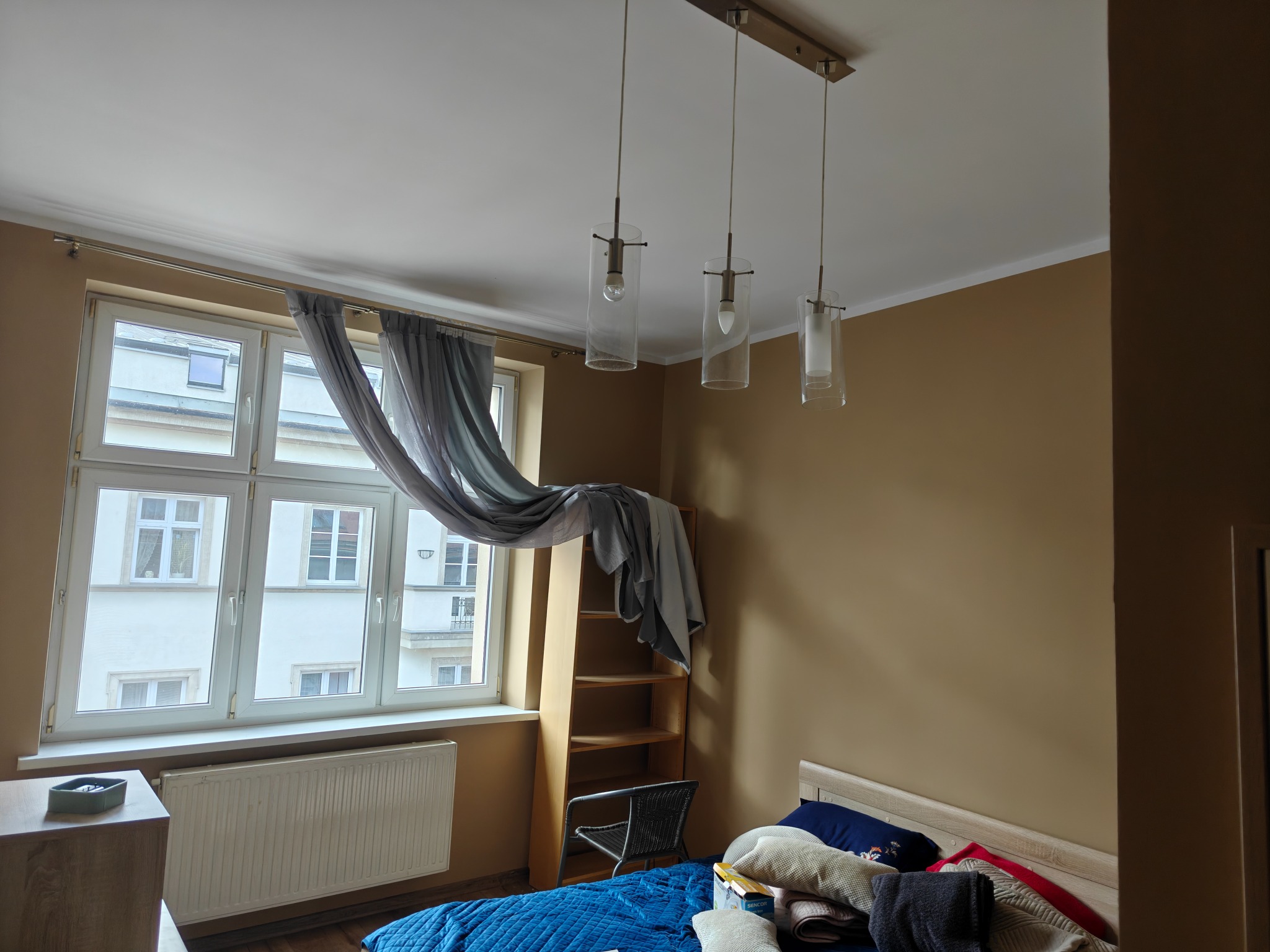 Pokój po malowaniu ścian na jednolity, beżowy kolor. Widoczne okno z zasłonami, łóżko z niebieską narzutą i lampy wiszące. Minimalistyczny wystrój wnętrza.