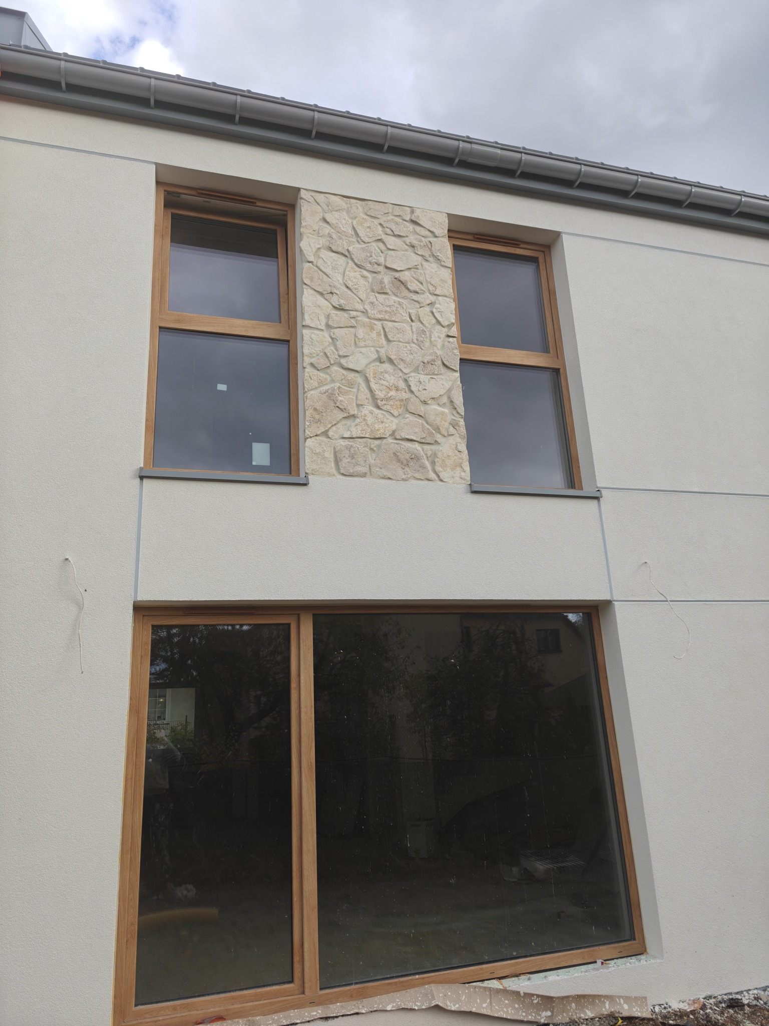 Fragment elewacji budynku z oknami w drewnianych ramach i dekoracyjnym panelem z kamienia. Świeżo otynkowana ściana w jasnym odcieniu. Widoczne ślady prac wykończeniowych.