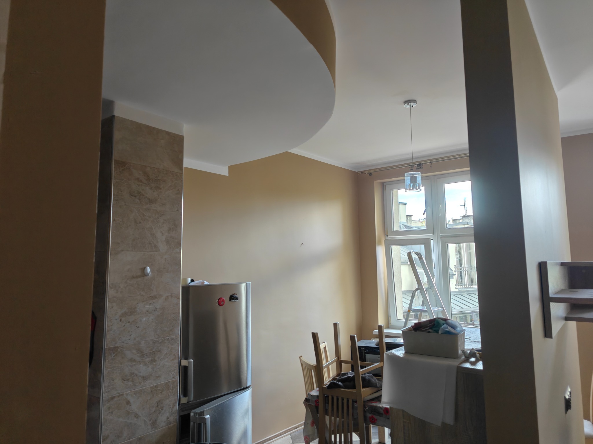 Beżowe ściany po malowaniu w mieszkaniu z zaokrąglonym sufitem, widoczne meble i drabina przy oknie. Ściana wykończona płytkami imitującymi marmur.