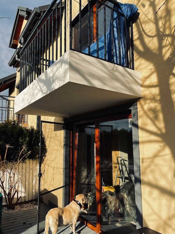 Żółta elewacja domu z balkonem i psem przy drzwiach tarasowych. Na balkonie widoczna plandeka. Cień drzewa na ścianie. Wnętrze w trakcie prac wykończeniowych.