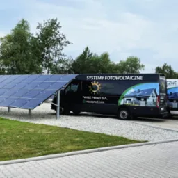 Rząd paneli słonecznych obok zaparkowanych samochodów dostawczych z logo firmy i hasłem 'Systemy Fotowoltaiczne'.
