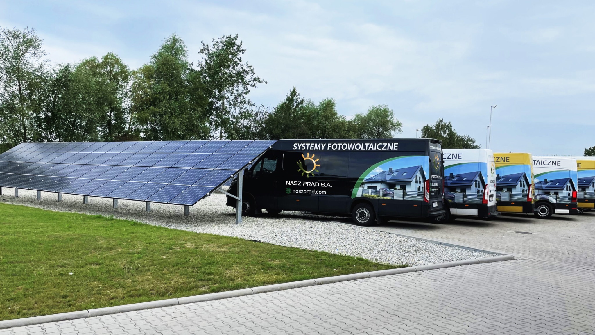 Rząd paneli słonecznych obok zaparkowanych samochodów dostawczych z logo firmy i hasłem 'Systemy Fotowoltaiczne'.