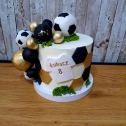 Tort urodzinowy z motywem piłkarskim: biały krem, czarno-złote heksagony, piłki i złote kule, personalizowany koszulką z imieniem Łukasz i numerem 8, na drewnianym stole i tle z desek.