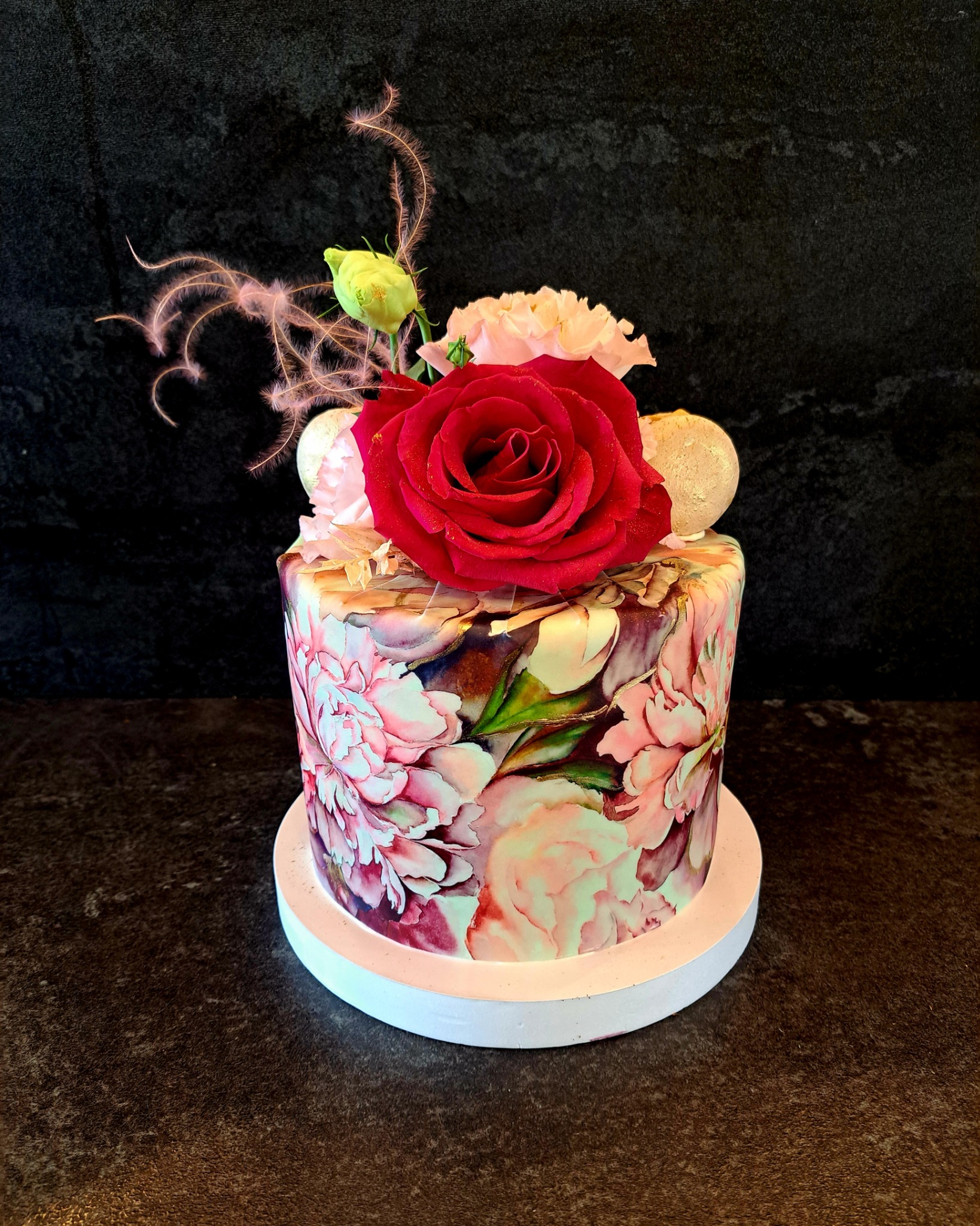 Tort artystyczny z motywem kwiatowym i czerwoną różą na wierzchu, ozdobiony białymi kulkami i delikatnymi piórkami, na białym podkładzie, na tle ciemnej powierzchni.