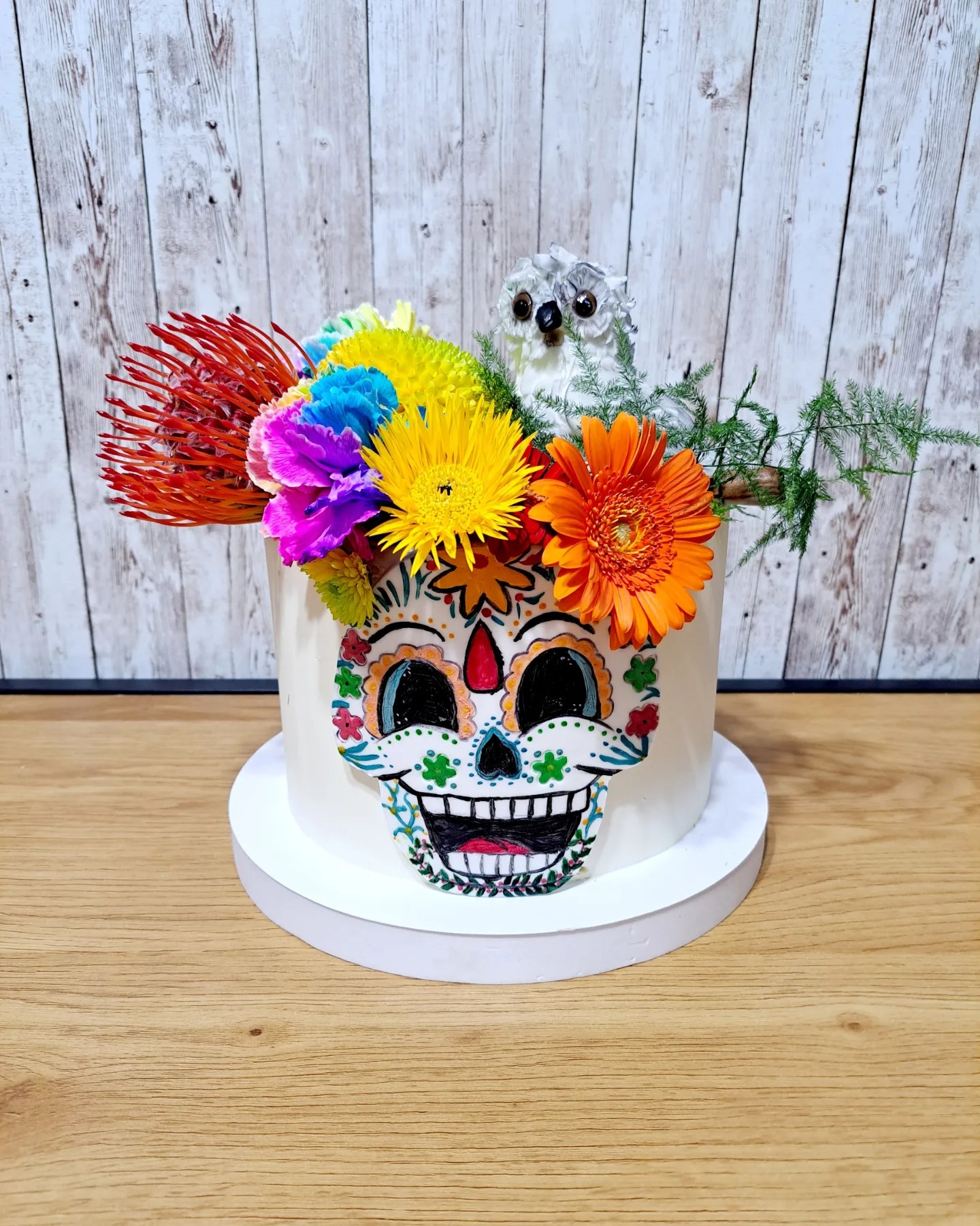 Tort zdobiony motywem czaszki z kolorowymi kwiatami i figurką sowy, na drewnianym blacie i tle z desek.