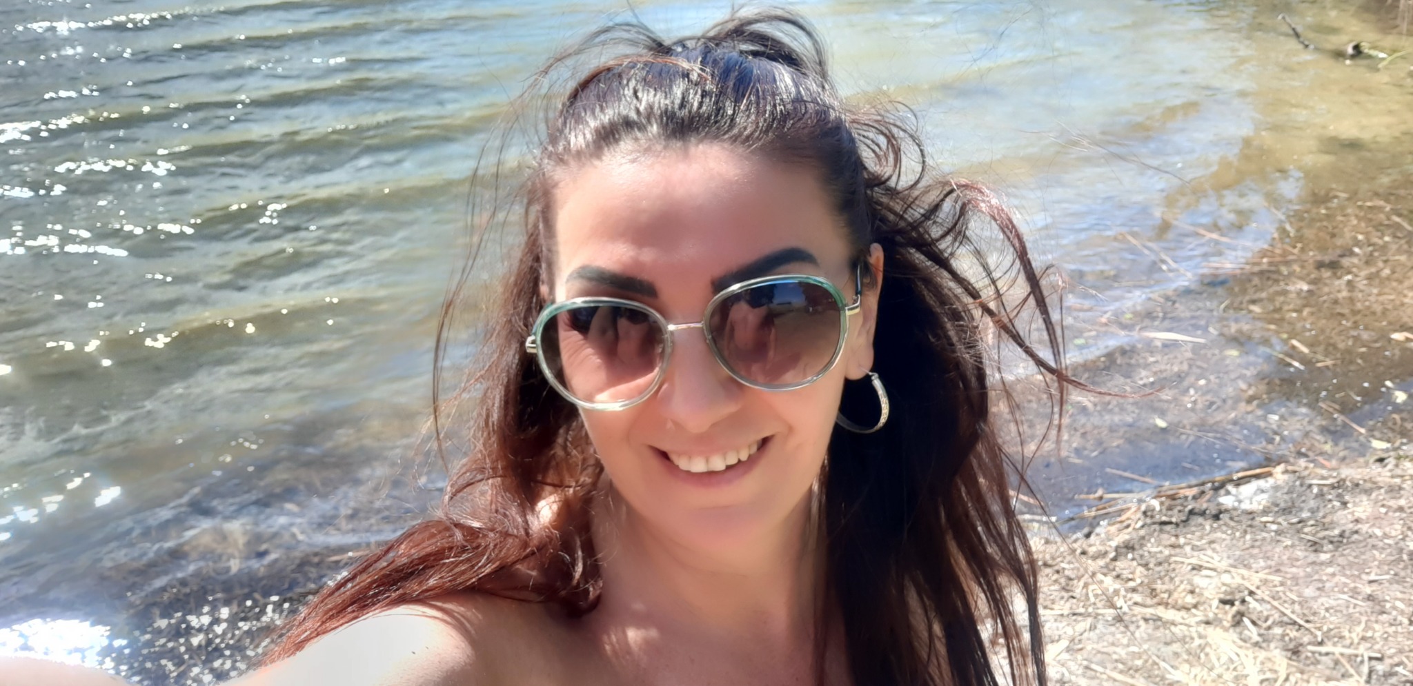 Uśmiechnięta kobieta w okularach przeciwsłonecznych z brązowymi włosami na tle jeziora w słoneczny dzień.