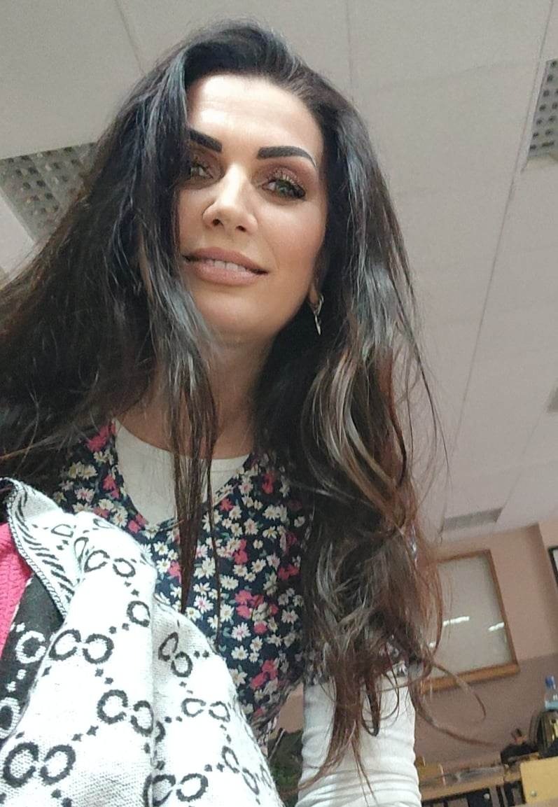 Portret kobiety z długimi, ciemnymi włosami z pasemkami, w kwiecistej bluzce, trzymającej wzorzysty szal. Ujęcie z dołu w jasnym pomieszczeniu z widocznym fragmentem sufitu i biurka.