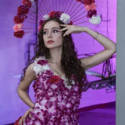 Kobieta z kręconymi włosami, ubrana w sukienkę i nakrycie głowy z żywych kwiatów, pozuje na tle fioletowej ściany z elementami konstrukcyjnymi.