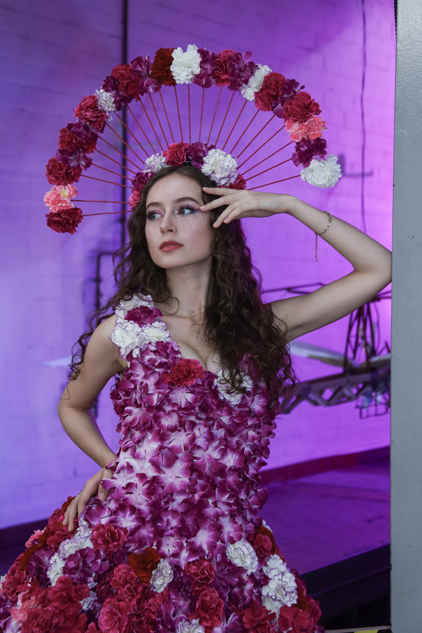 Kobieta z kręconymi włosami, ubrana w sukienkę i nakrycie głowy z żywych kwiatów, pozuje na tle fioletowej ściany z elementami konstrukcyjnymi.