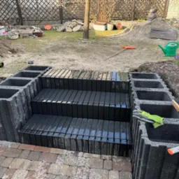 Budowa schodów z betonowych bloczków na zewnątrz, widoczne narzędzia i kostka brukowa. Etap prac w ogrodzie, w tle trawa i roślinność.
