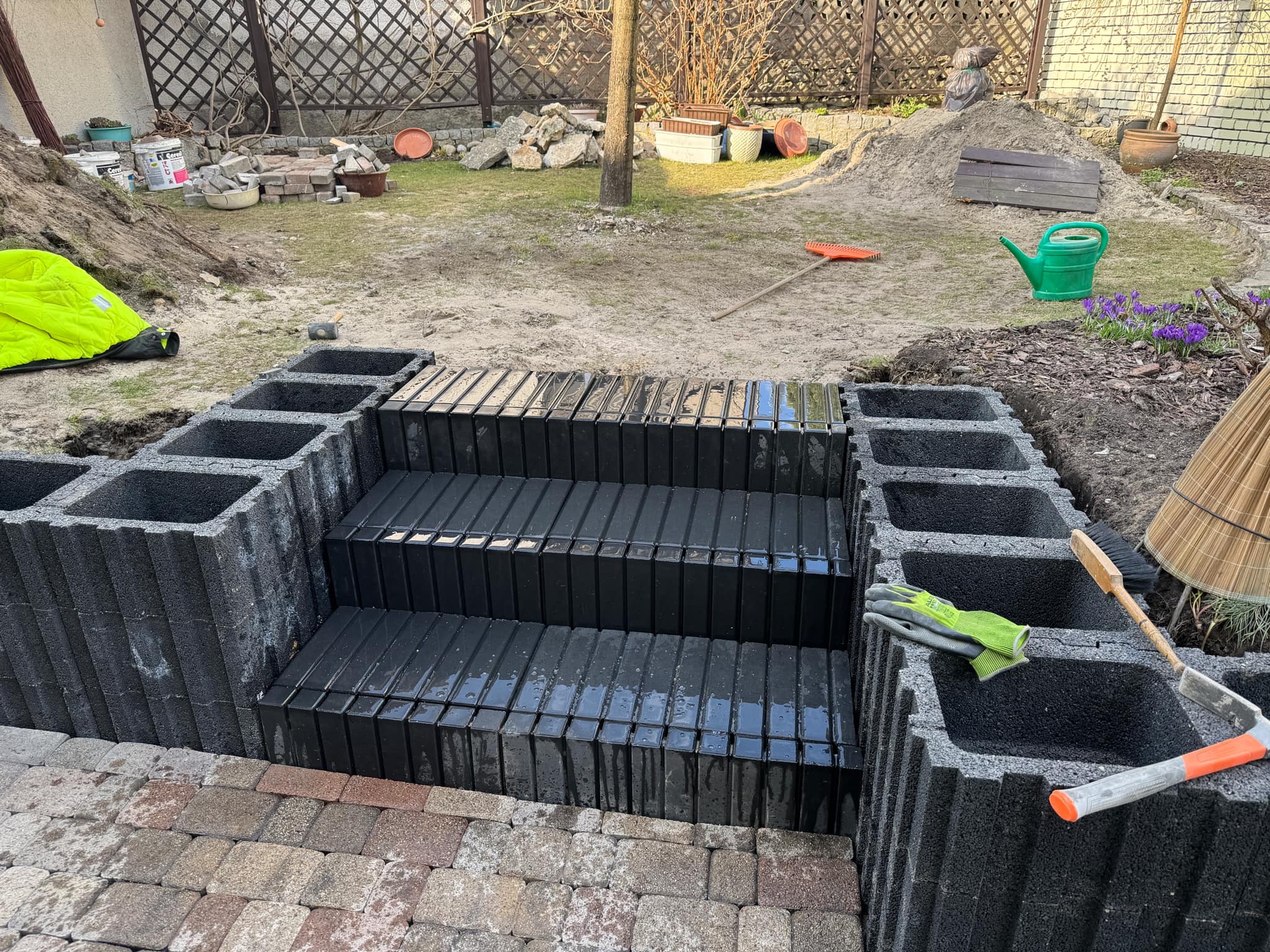 Budowa schodów z betonowych bloczków na zewnątrz, widoczne narzędzia i kostka brukowa. Etap prac w ogrodzie, w tle trawa i roślinność.