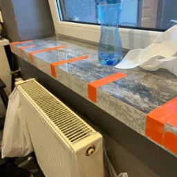 Marmurowy parapet zabezpieczony pomarańczową taśmą malarską przed zabrudzeniem podczas malowania ściany, obok grzejnik i butelka wody mineralnej.