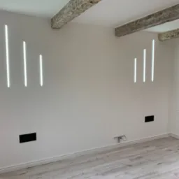 Nowoczesne oświetlenie ścienne LED wpuszczone w ścianę, z dekoracyjnymi belkami stropowymi i jasną podłogą imitującą drewno w wykończonym wnętrzu.