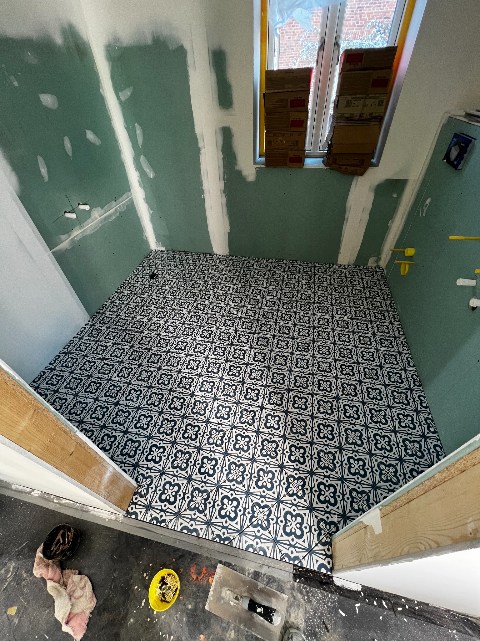 Łazienka w trakcie remontu: ułożona podłoga z wzorzystych płytek, ściany przygotowane do malowania z widocznymi punktami elektrycznymi, okno zasłonięte kartonami, narzędzia i akcesoria na podłodze.