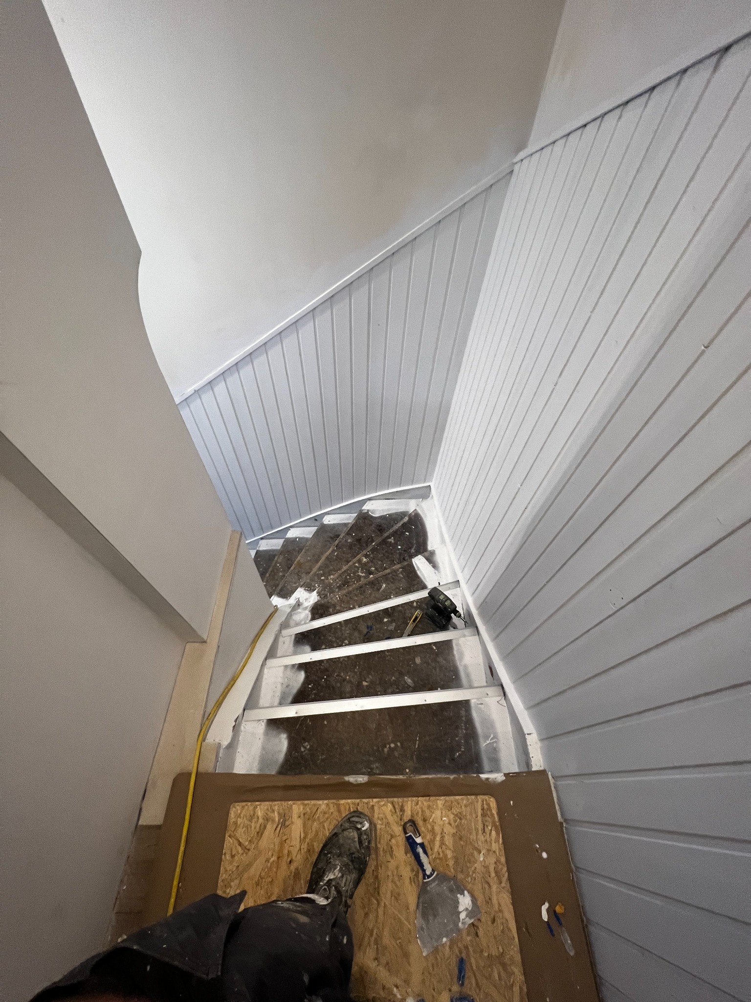 Widok z góry na klatkę schodową w trakcie remontu; widoczne szare panele ścienne, częściowo pomalowane schody, narzędzia i stopa w roboczym bucie na płycie OSB.