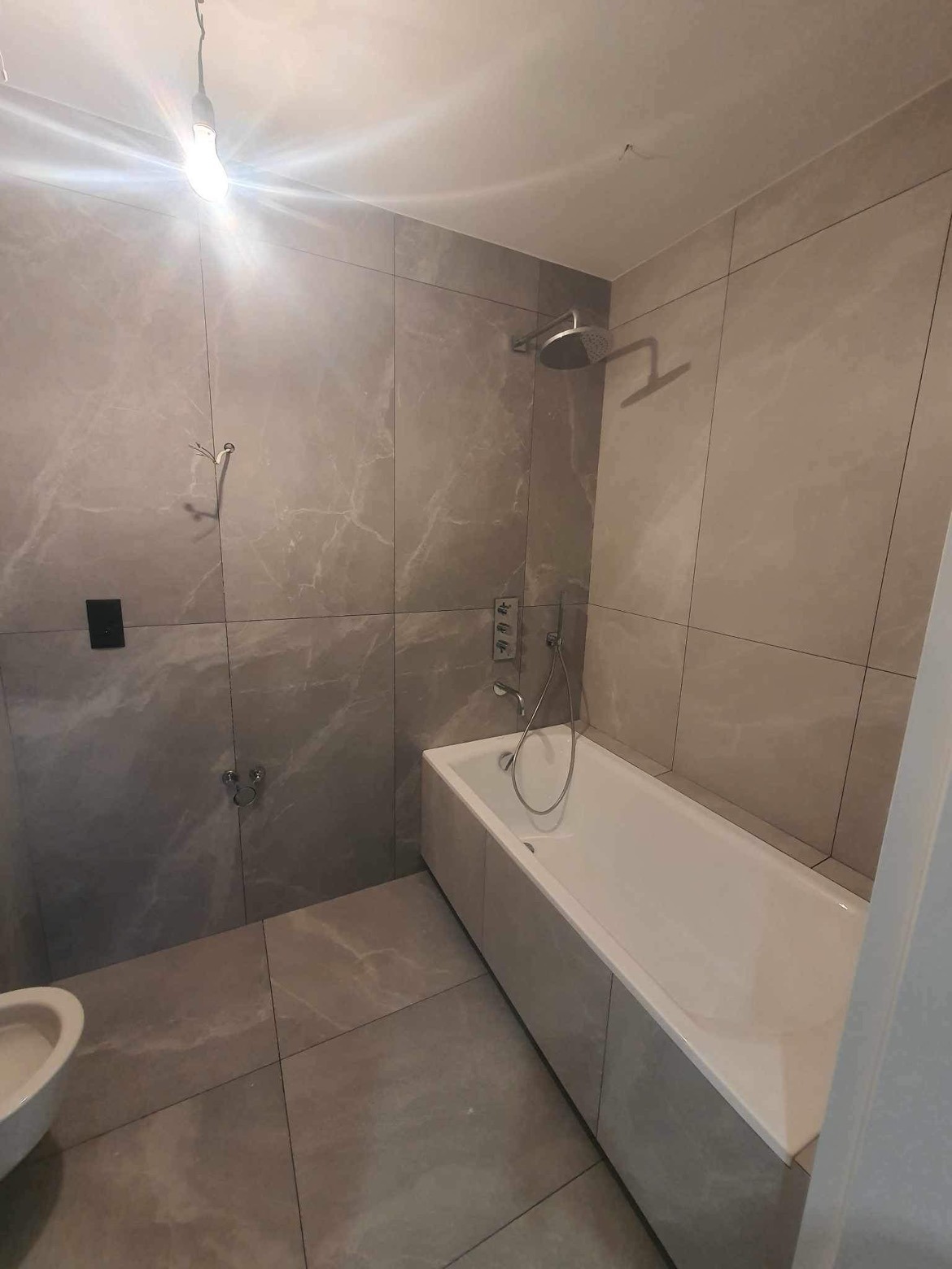 Wykończona łazienka z wanną obudowaną płytkami, szare płytki imitujące marmur na ścianach i podłodze, deszczownica i bateria podtynkowa, widoczny fragment toalety.