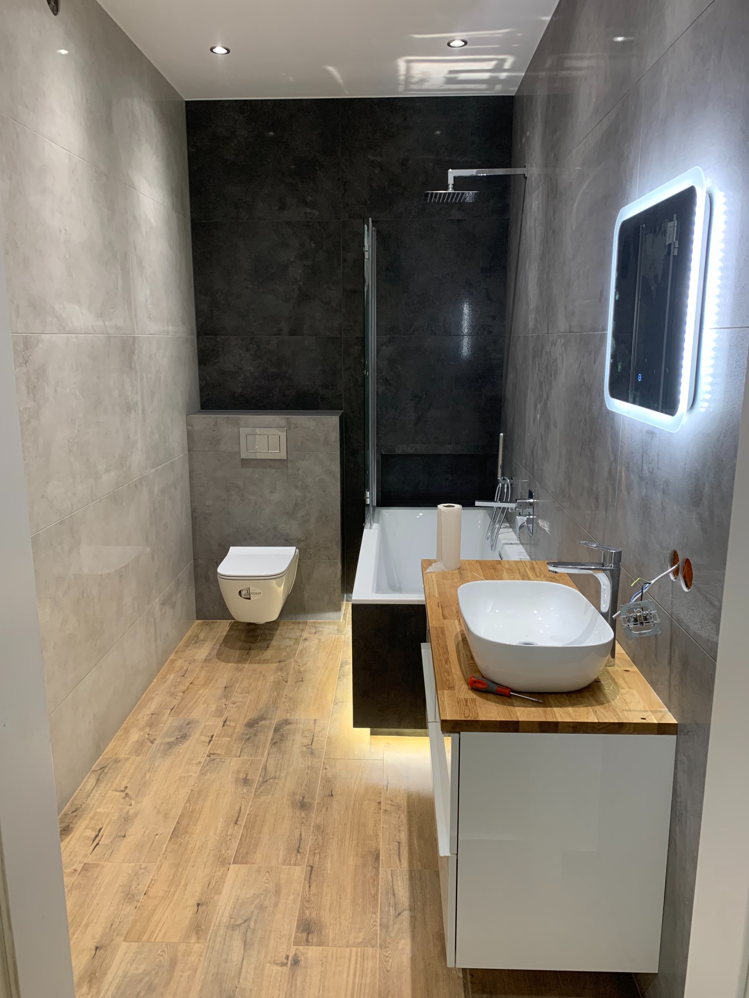 Nowoczesna łazienka z drewnopodobną podłogą, szarymi płytkami na ścianach, podświetlanym lustrem i umywalką nablatową z drewnianym blatem; widoczny etap montażu elektryki i śrubokręt na blacie.