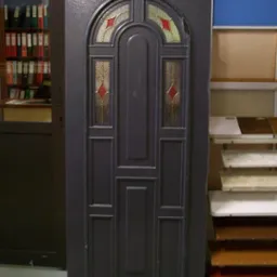 Szare drzwi zewnętrzne z dekoracyjnymi szybkami z motywem rombów w czerwonej tonacji, eksponowane w sklepie z materiałami budowlanymi.