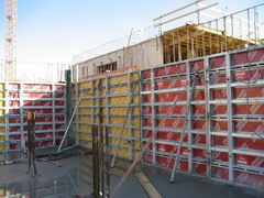 Szalunki systemowe ścian z wypełnieniem w kolorze czerwonym i żółtym, wsparte na metalowych podporach, widoczne pręty zbrojeniowe i świeżo wylany beton w wykopie fundamentowym, w tle konstrukcja...