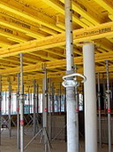 Widok na konstrukcję szalunkową stropową z podporami teleskopowymi, widoczne żółte płyty szalunkowe i stalowe podpory, ujęcie z dołu.