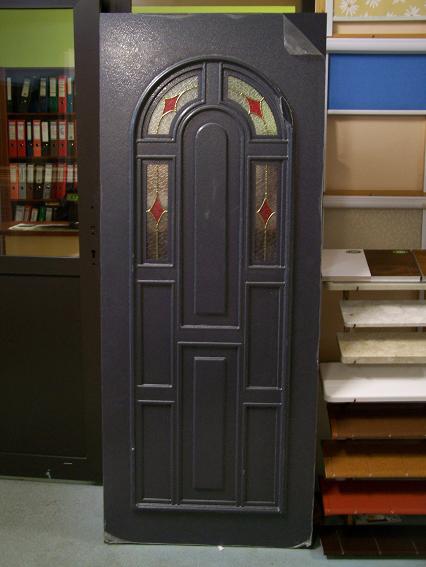 Szare drzwi zewnętrzne z dekoracyjnymi szybkami z motywem rombów w czerwonej tonacji, eksponowane w sklepie z materiałami budowlanymi.