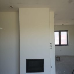 Filip Kowalski - Wnętrze w trakcie wykańczania z zabudową kominkową z płyt gipsowo-kartonowych, widoczne szpachlowane ściany i sufit z widocznymi niedoskonałościami przed malowaniem, okno zabezpieczone folią malarską.