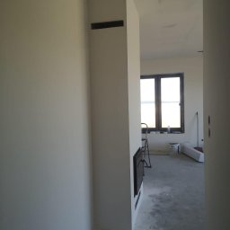 Filip Kowalski - Wnętrze w trakcie wykańczania: świeżo pomalowane ściany w odcieniach bieli, widoczne ślady po pracach na podłodze, okno z widokiem na zewnątrz i drabina malarska w tle.