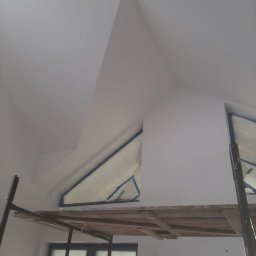 Filip Kowalski - Świeżo pomalowane poddasze ze skośnym sufitem i oknem zabezpieczonym folią malarską, widoczne rusztowanie.