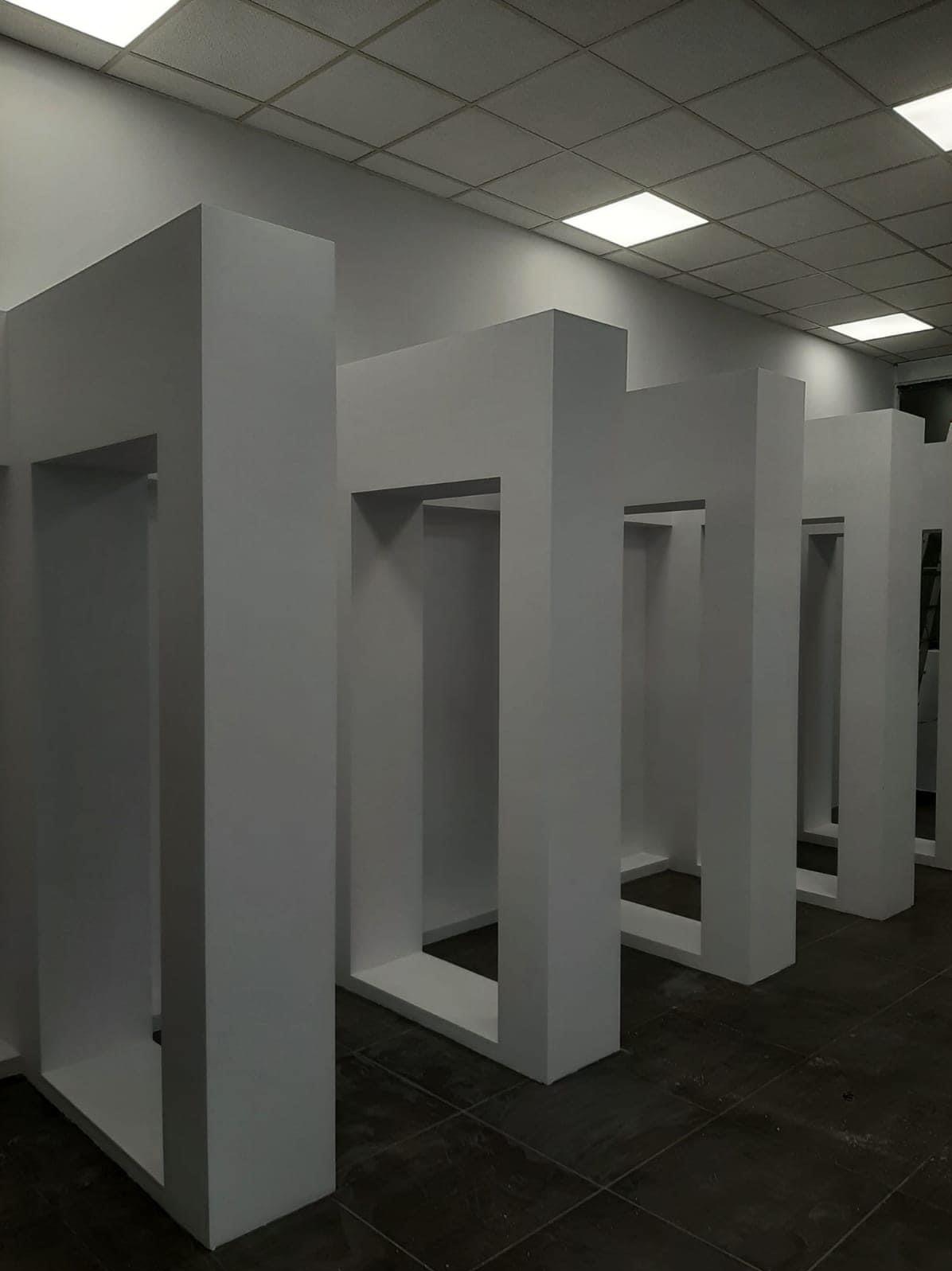 Białe, prostopadłościenne konstrukcje z płyt karton-gips, tworzące rzędy otwartych ram w jasnym pomieszczeniu z kasetonowym sufitem i ciemną podłogą.