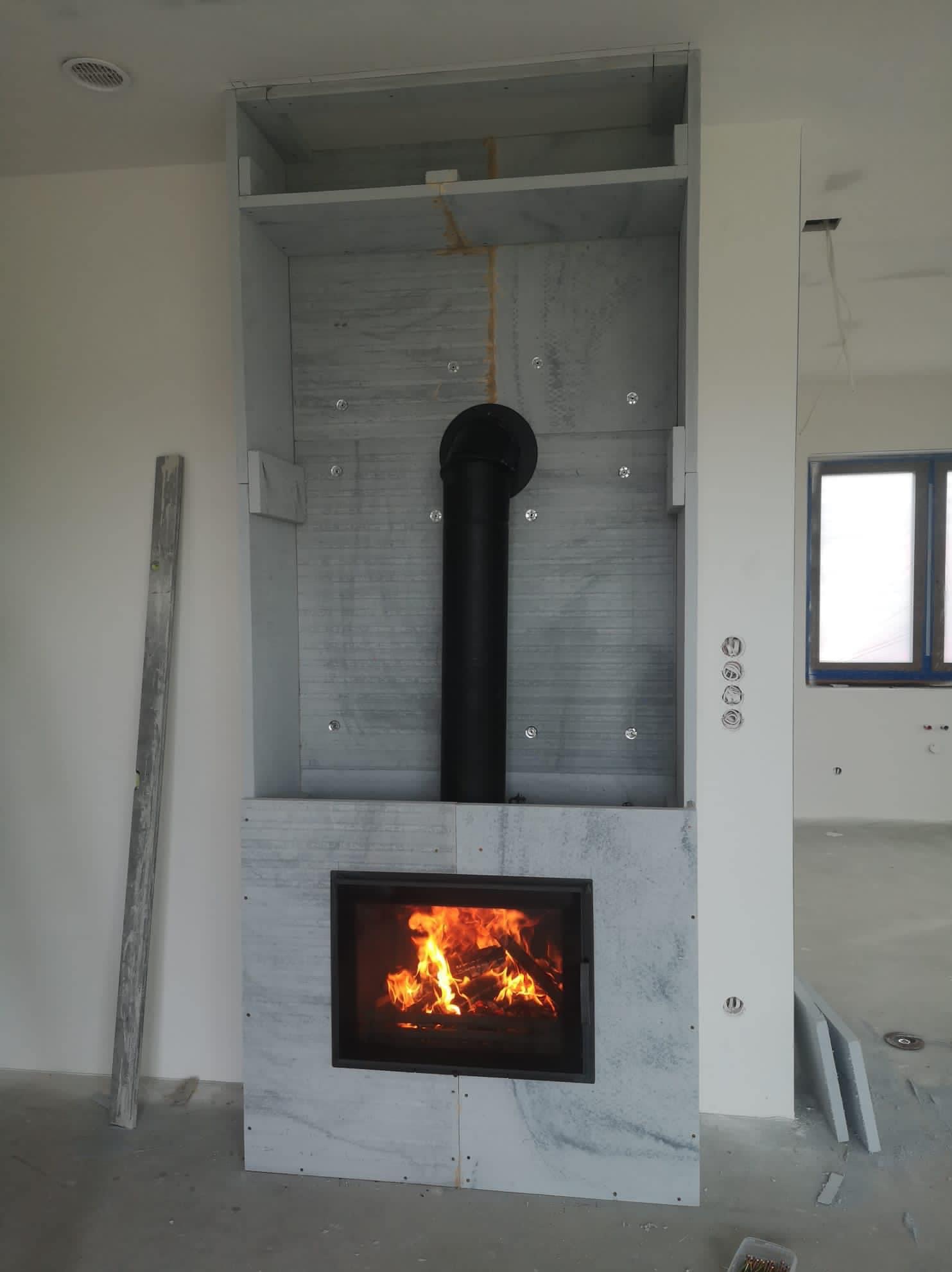 Wnętrze w trakcie wykańczania z zabudowanym kominkiem z czarnym wkładem, obudowanym płytami imitującymi marmur. Widoczny czarny przewód kominowy i poziomica oparta o ścianę. W tle okno...