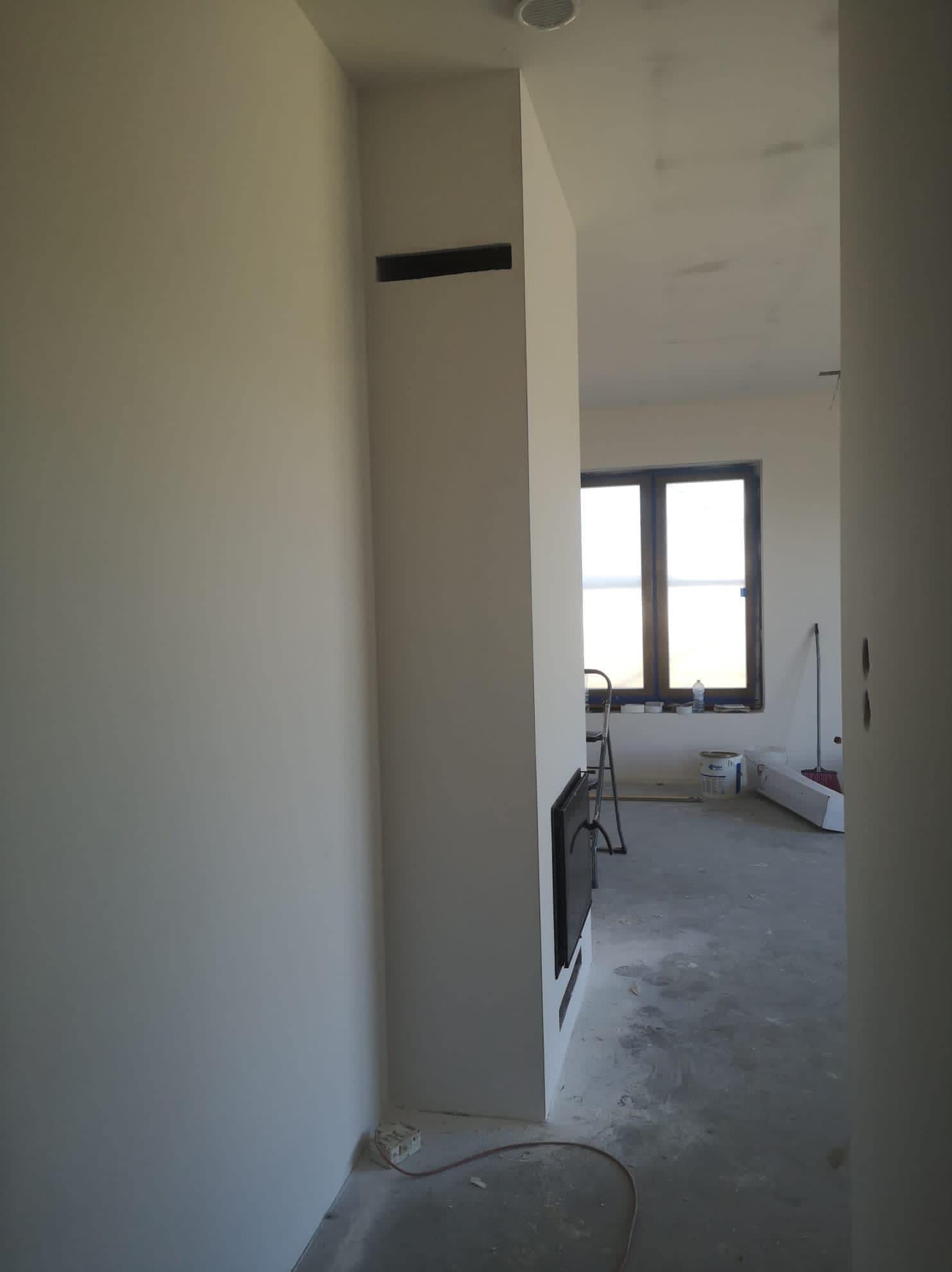 Wnętrze w trakcie wykańczania: świeżo pomalowane ściany w odcieniach bieli, widoczne ślady po pracach na podłodze, okno z widokiem na zewnątrz i drabina malarska w tle.