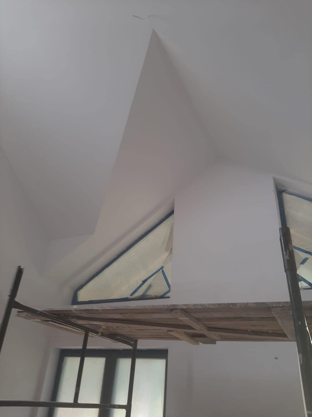 Świeżo pomalowane poddasze ze skośnym sufitem i oknem zabezpieczonym folią malarską, widoczne rusztowanie.