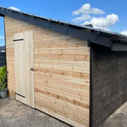 Nowo wybudowana drewniana konstrukcja ogrodowa z jasnego drewna, z pojedynczymi drzwiami i czarnym wykończeniem dachu, widoczna z zewnątrz, obok rosną tuje, a na ziemi leży łopata i wiadro.