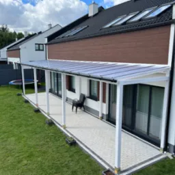 Nowoczesna biała konstrukcja z przezroczystym zadaszeniem przylegająca do domu, z wyłożonym kostką brukową tarasem, trawnikiem i stosem krzeseł w tle.