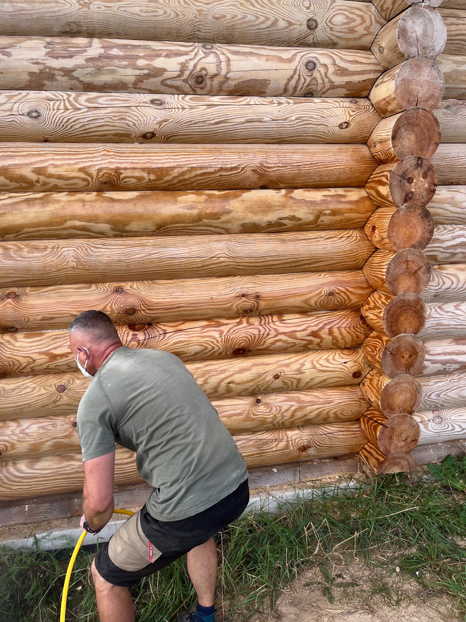 Pracownik w masce ochronnej czyści drewnianą ścianę domu z bali za pomocą węża z wodą, widoczne świeżo impregnowane drewno.