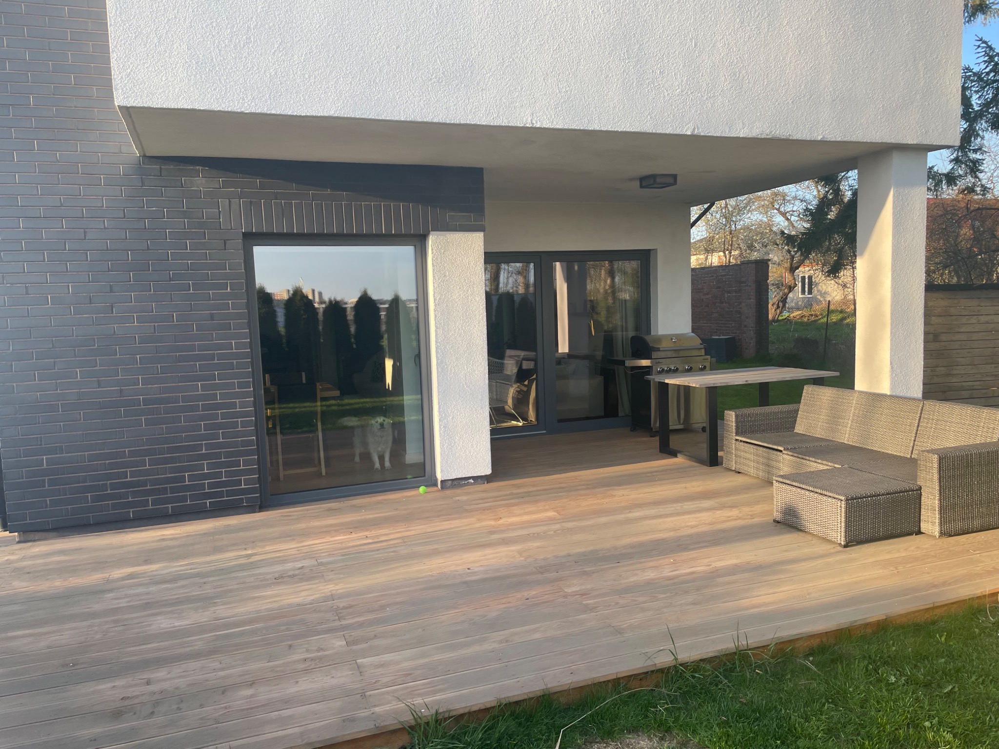 Nowoczesny dom z drewnianym tarasem, szarym ceglanym wykończeniem i meblami ogrodowymi z rattanu, widok na psa w oknie