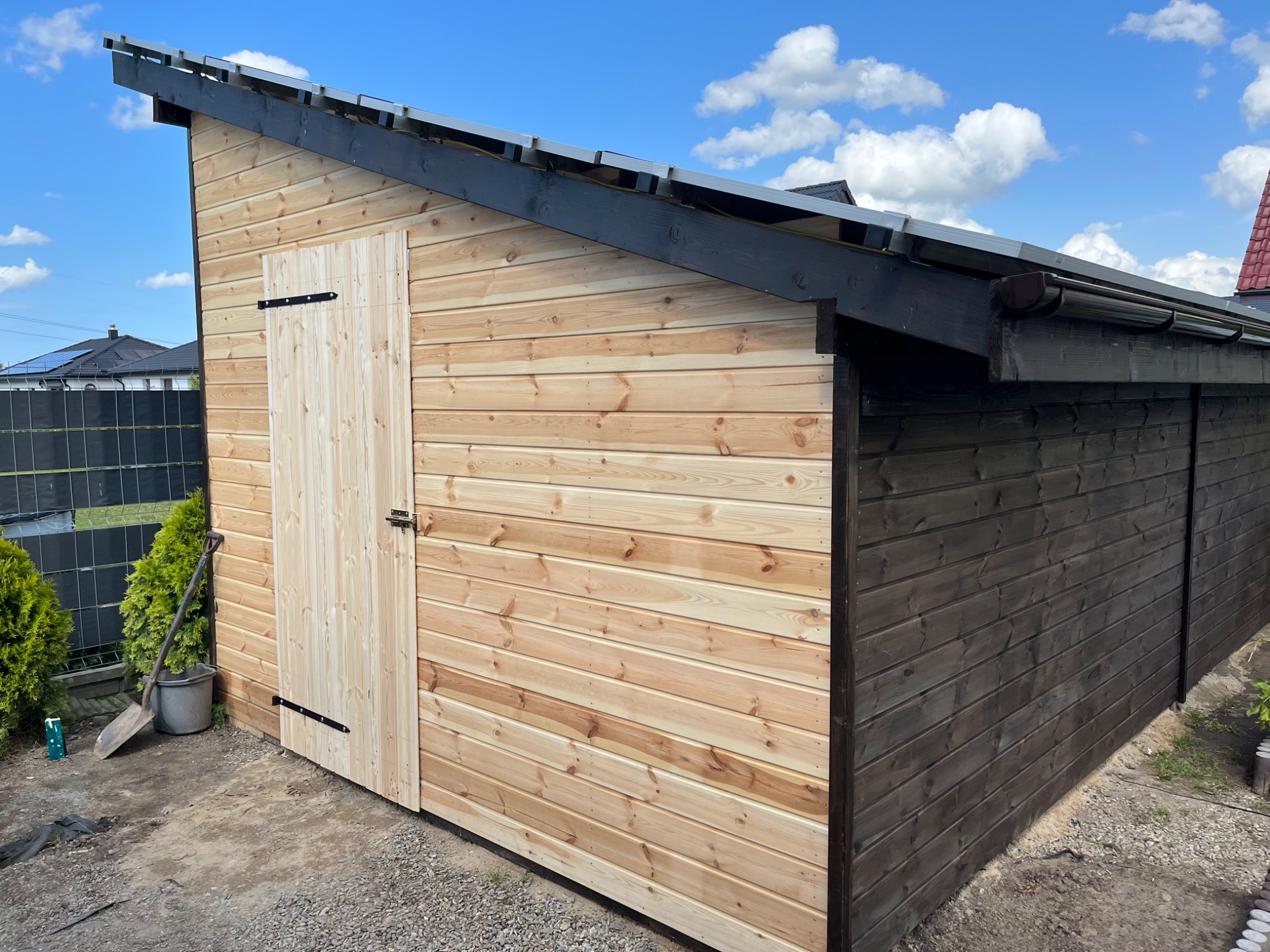 Nowo wybudowana drewniana konstrukcja ogrodowa z jasnego drewna, z pojedynczymi drzwiami i czarnym wykończeniem dachu, widoczna z zewnątrz, obok rosną tuje, a na ziemi leży łopata i wiadro.