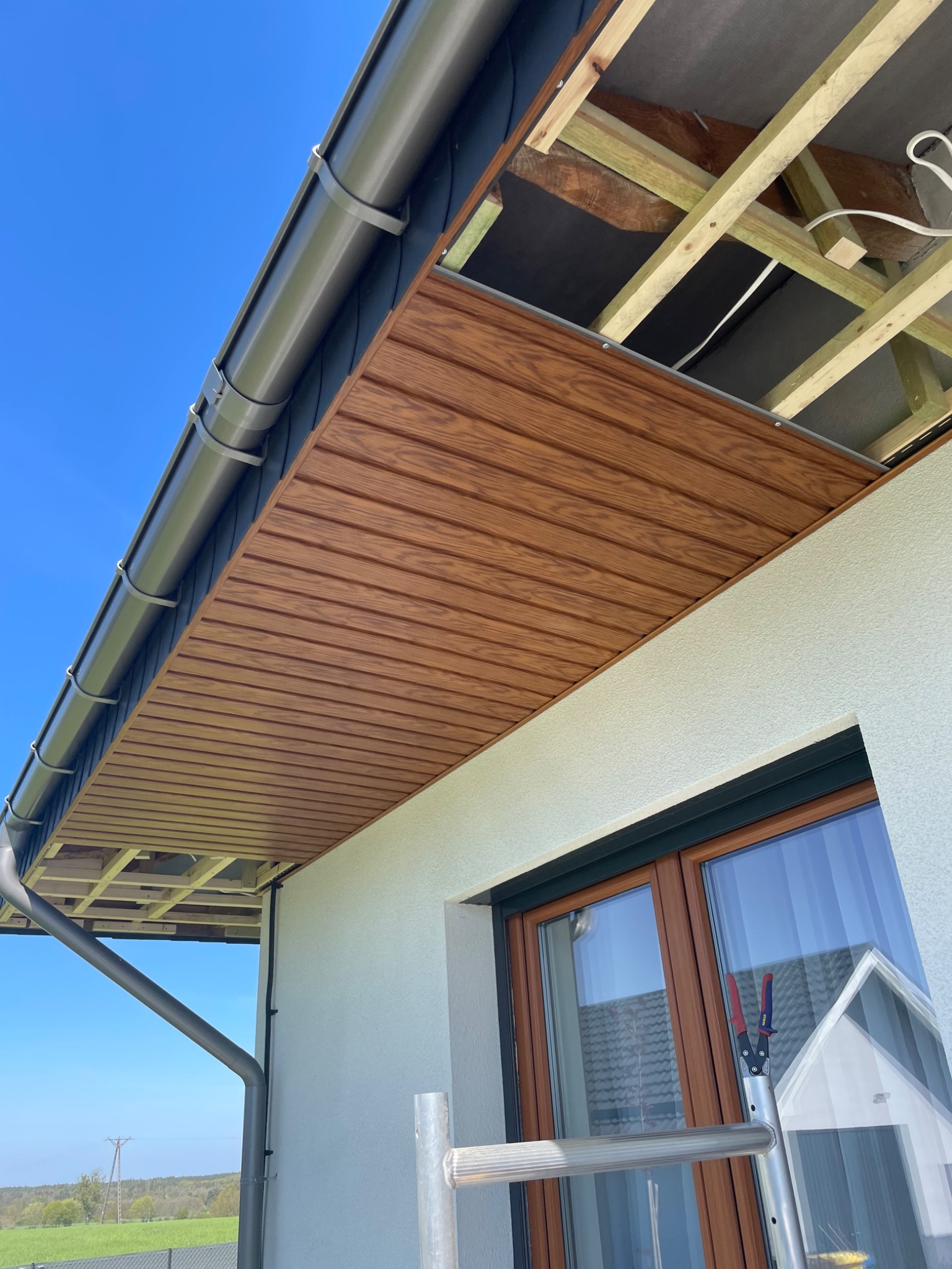 Montaż podbitki dachowej z paneli imitujących drewno, widoczne krokwie i orynnowanie, fragment drabiny oparty o okno.