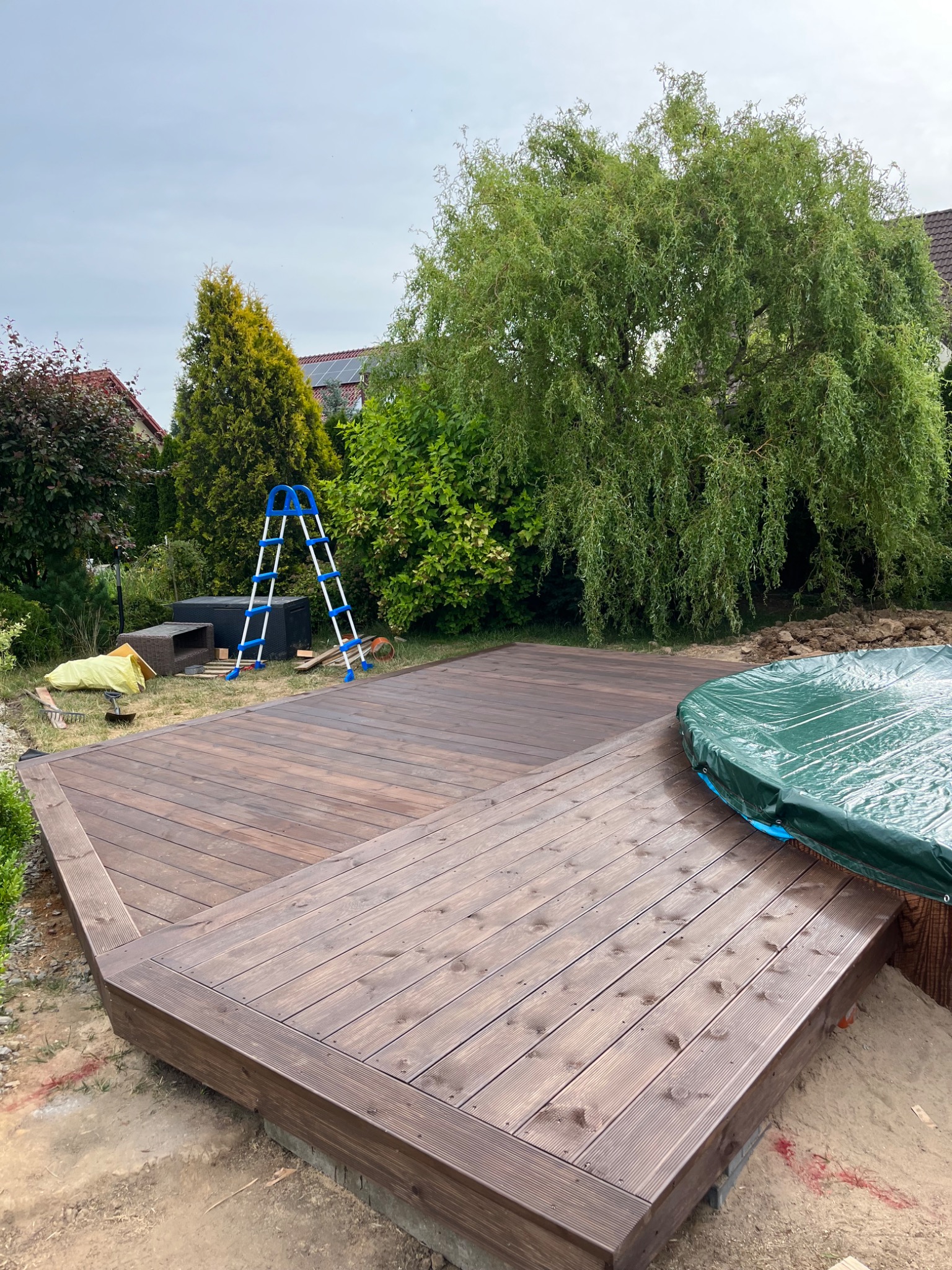 Nowo wybudowany, ciemnobrązowy taras z drewna w ogrodzie, z widocznymi schodkami, częściowo przykryty basen z zieloną plandeką i drabina w tle.