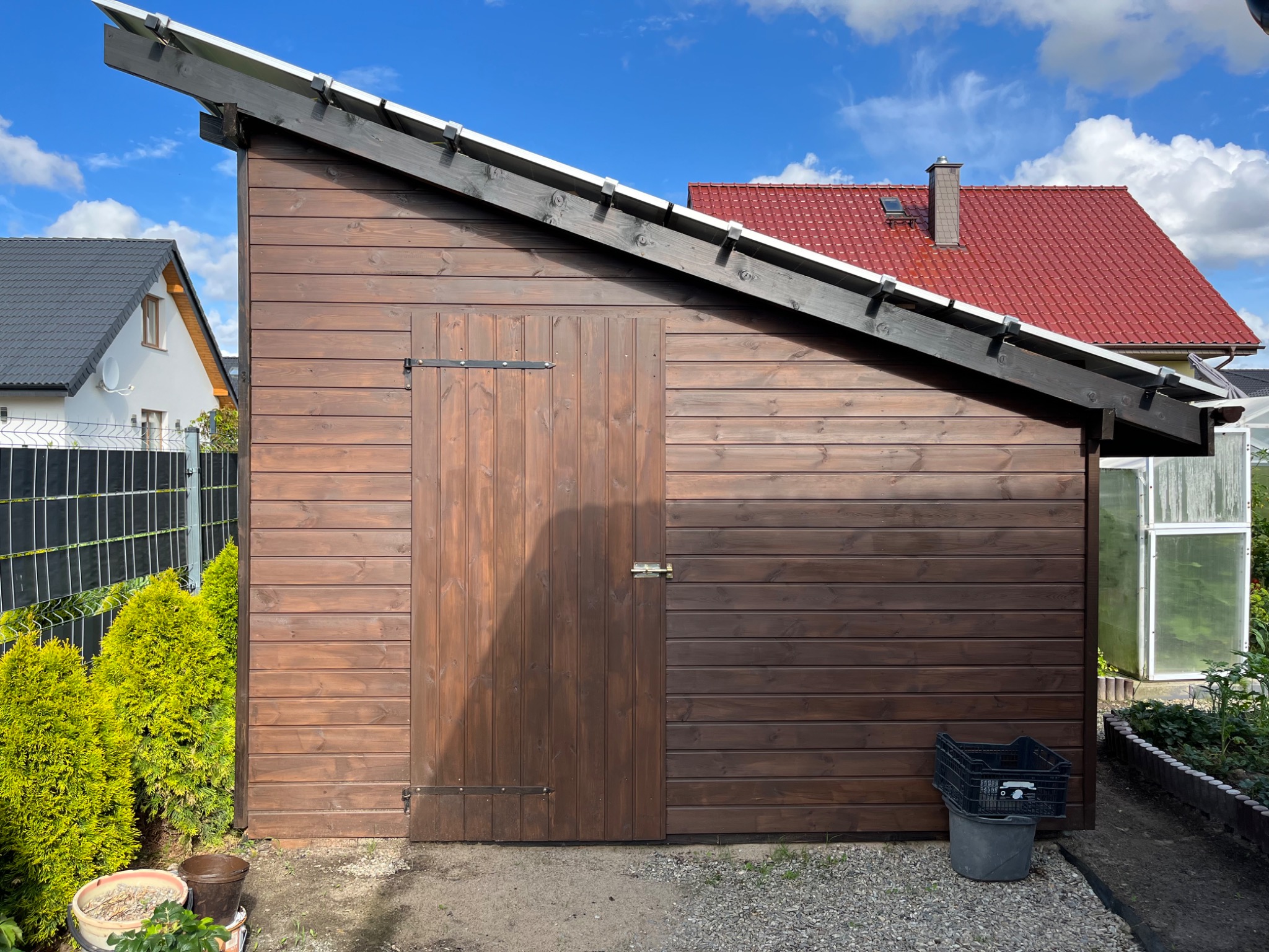 Brązowa drewniana szopa ogrodowa z jednospadowym dachem i drzwiami na metalowych zawiasach, obok zielone tuje i skrzynka na wiadro, w tle domy z czerwoną i szarą dachówką.