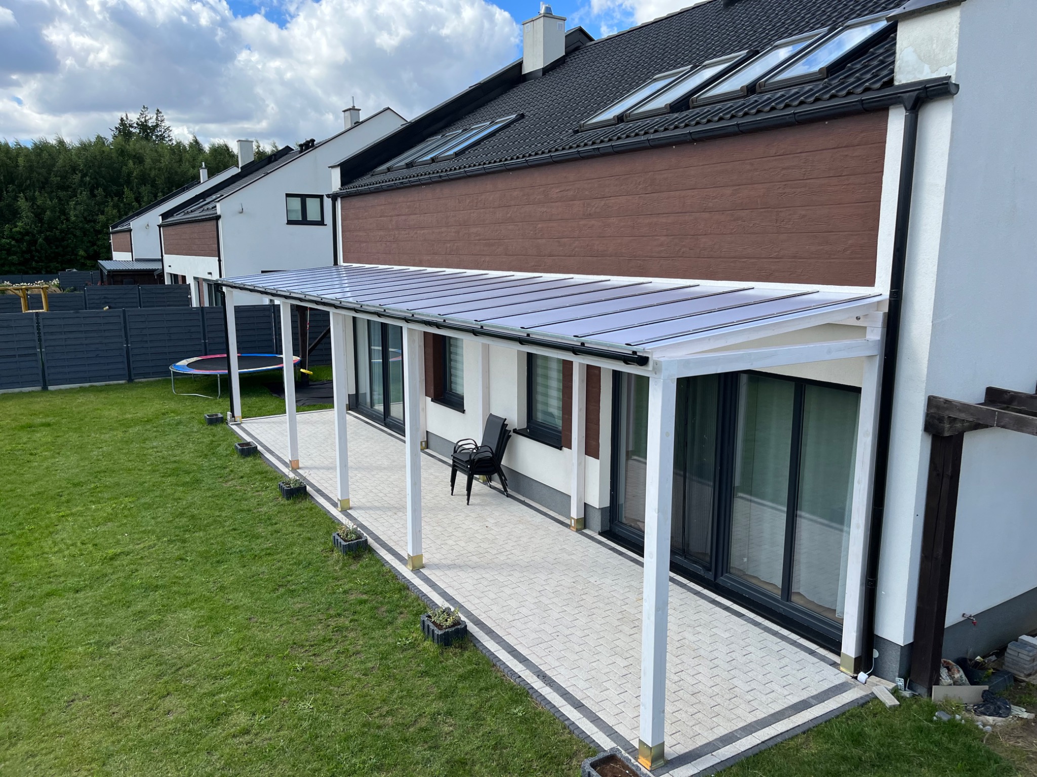 Nowoczesna biała konstrukcja z przezroczystym zadaszeniem przylegająca do domu, z wyłożonym kostką brukową tarasem, trawnikiem i stosem krzeseł w tle.