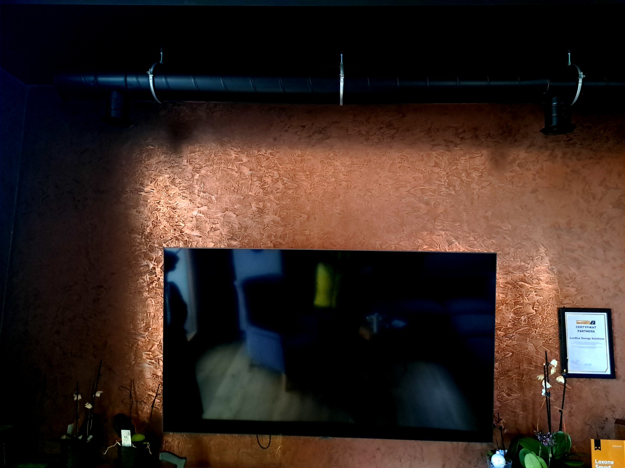 Dekoracyjna ściana w kolorze miedzianym z efektem strukturalnym, widoczna rura wentylacyjna pod sufitem, telewizor na ścianie, certyfikat partnerski powieszony obok, doniczki z roślinami na dole.