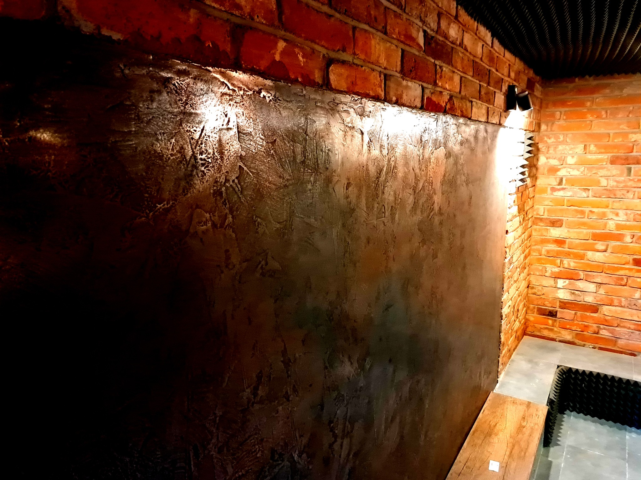 Dekoracyjna ściana z efektem betonu architektonicznego w pomieszczeniu z ceglanymi ścianami i oświetleniem punktowym.