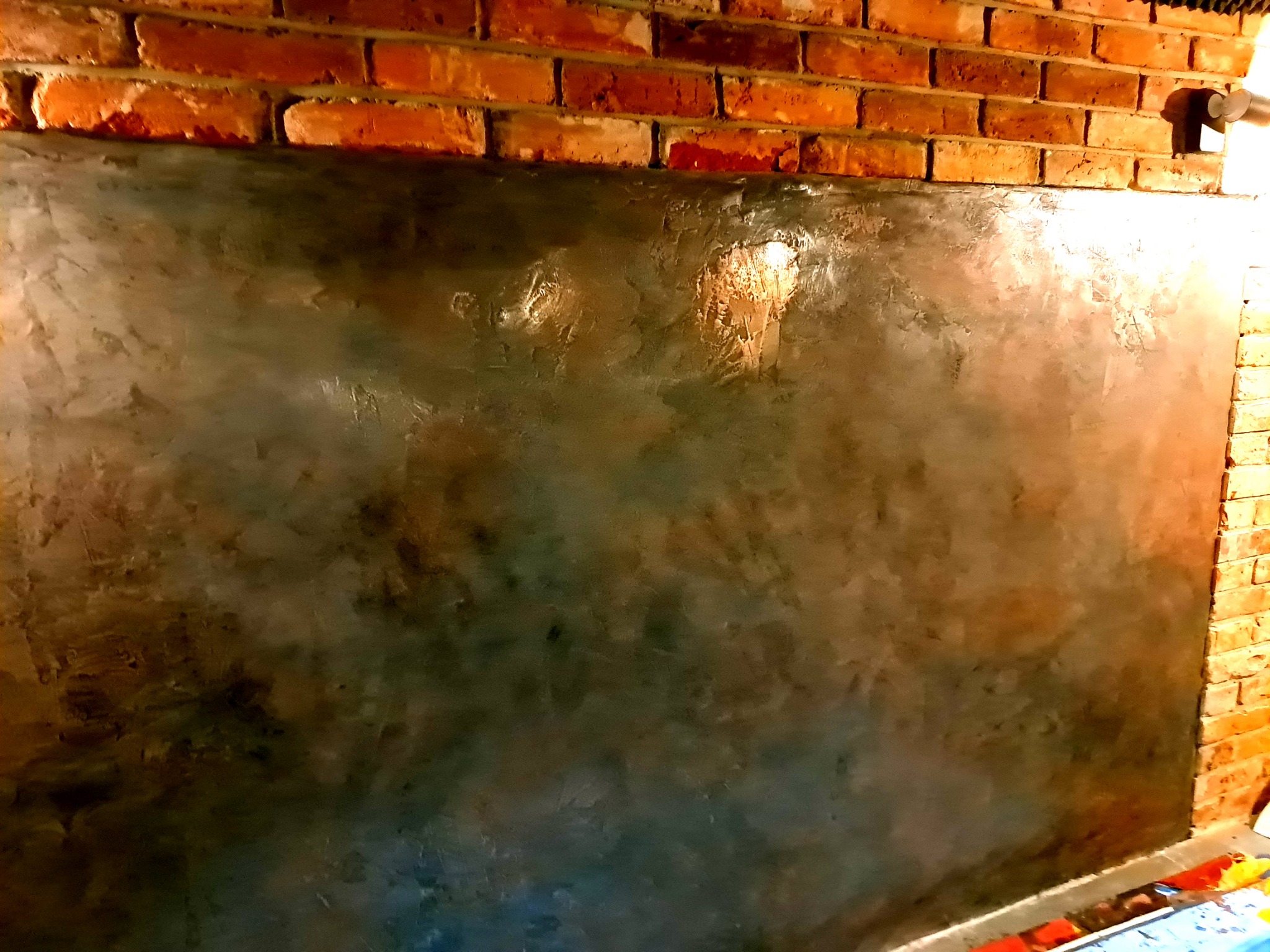 Ściana z efektem betonu architektonicznego, górna część wykończona czerwoną cegłą, widoczne ślady po szpachlowaniu i polerowaniu, ciepłe oświetlenie.