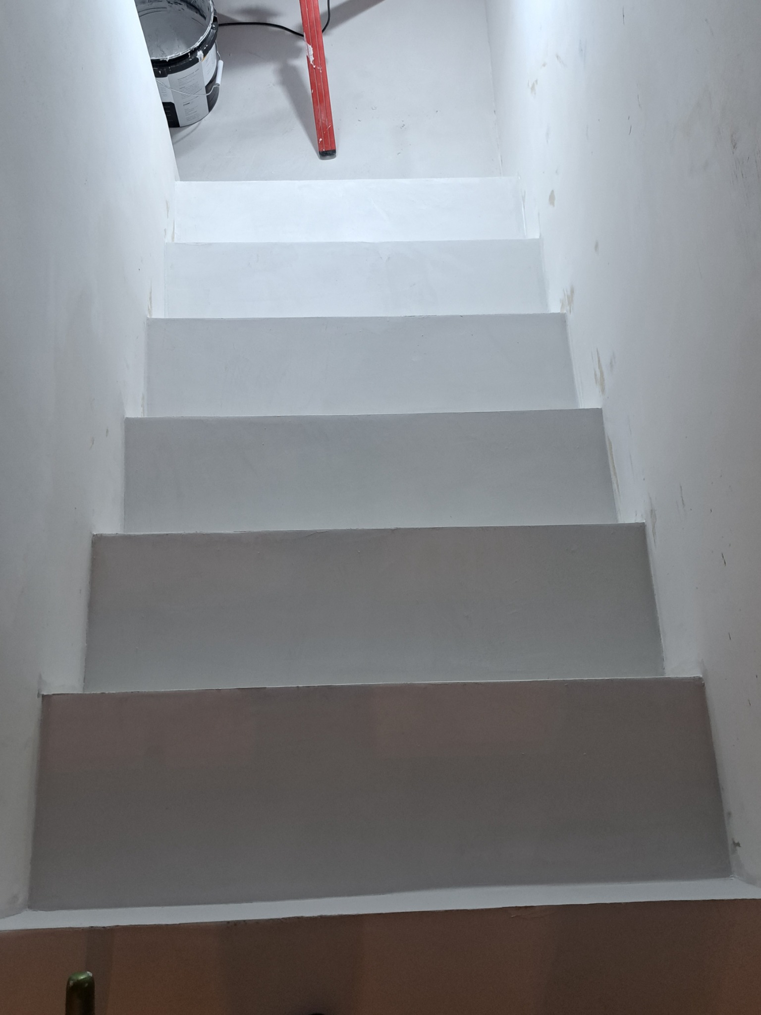 Pomieszczenie w trakcie malowania: białe schody i ściany, widoczne ślady farby, drabina i wiadro z farbą w tle.