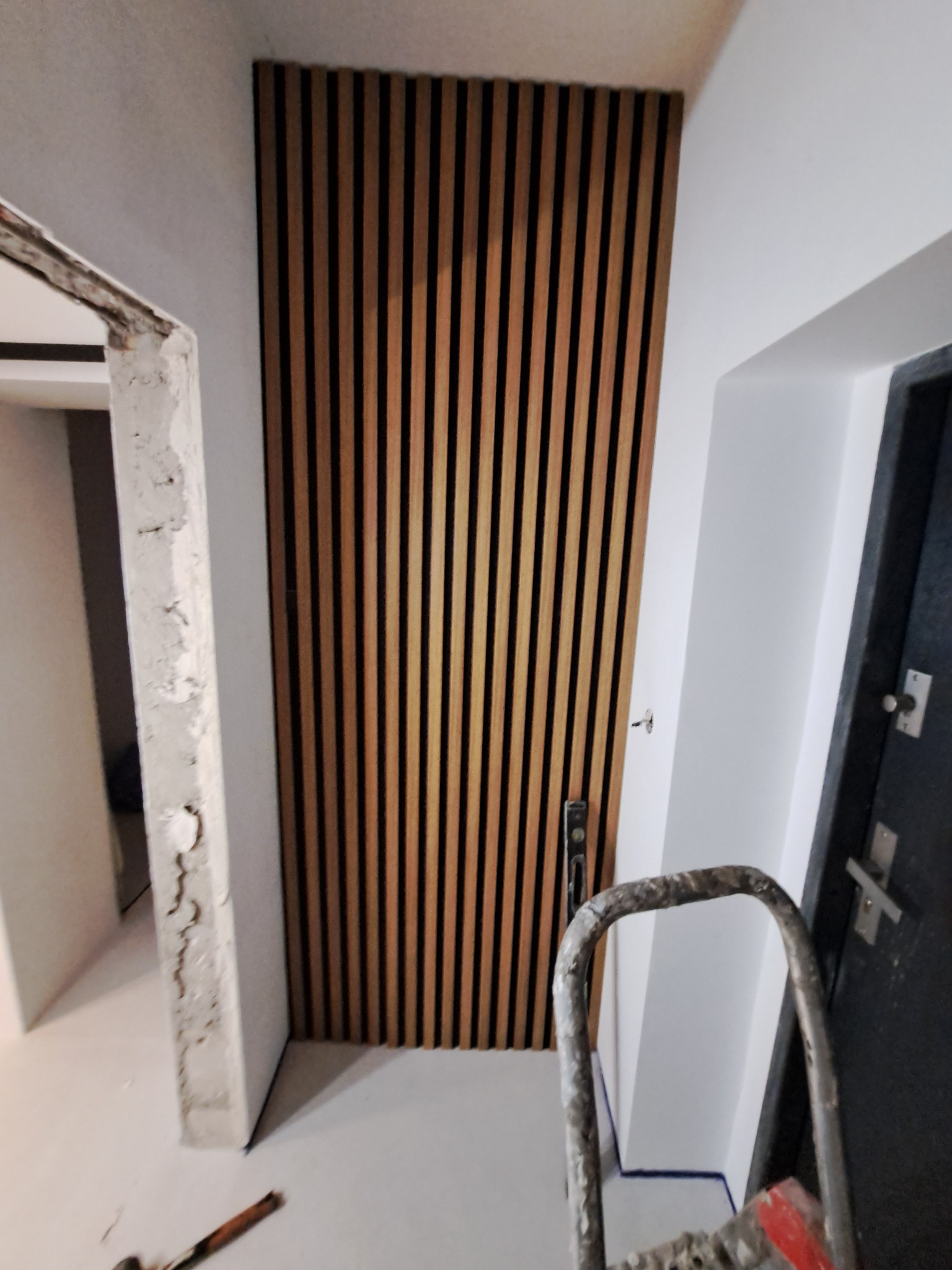 Montaż drewnianych lameli na ścianie w przedpokoju, widoczny fragment ościeżnicy w stanie surowym i drabina malarska.