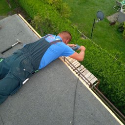 Usługi ciesielskie i wykończeniowe Wojciech Lesner - Pracownik w niebieskiej koszulce i granatowym kombinezonie mocuje drewniane elementy konstrukcji na dachu pokrytym papą, widok z góry na zielony ogród z krzewami i satelitą.