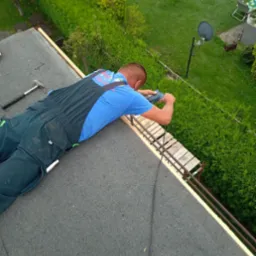 Pracownik w niebieskiej koszulce i granatowym kombinezonie mocuje drewniane elementy konstrukcji na dachu pokrytym papą, widok z góry na zielony ogród z krzewami i satelitą.